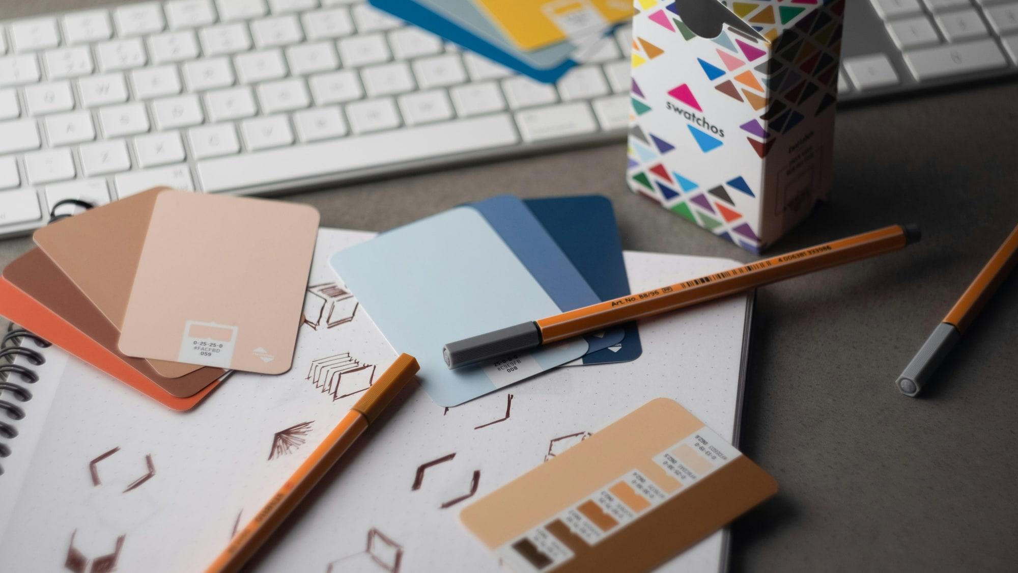 A designer's desk with colour swatch cards fanned across a sketchbook of logo marks, pencils beside them.