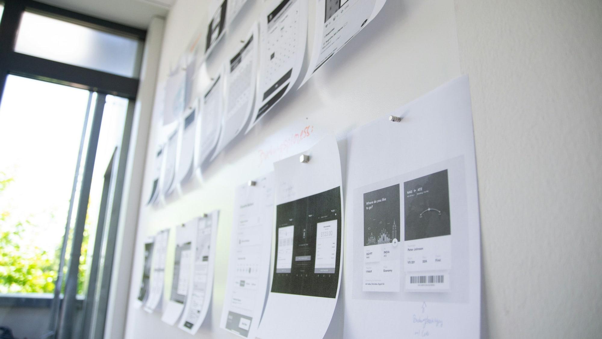 Printed page layouts pinned in a row along a white studio wall beside a window, each sheet a different screen of the same design, several marked up by hand.